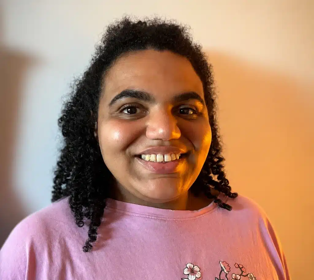 Portrait de Naomi, une jeune fille souriante dans sa vingtaine. Elle porte un t-shirt rose et s'est attachée les cheveux en demi queue.